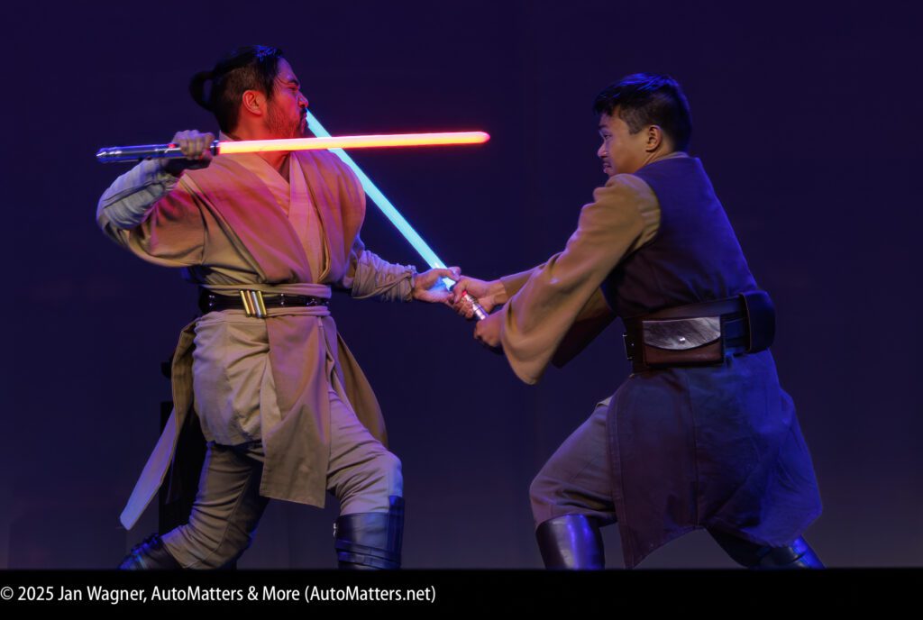 Two people dressed as Jedi duel with glowing lightsabers on stage against a purple background.