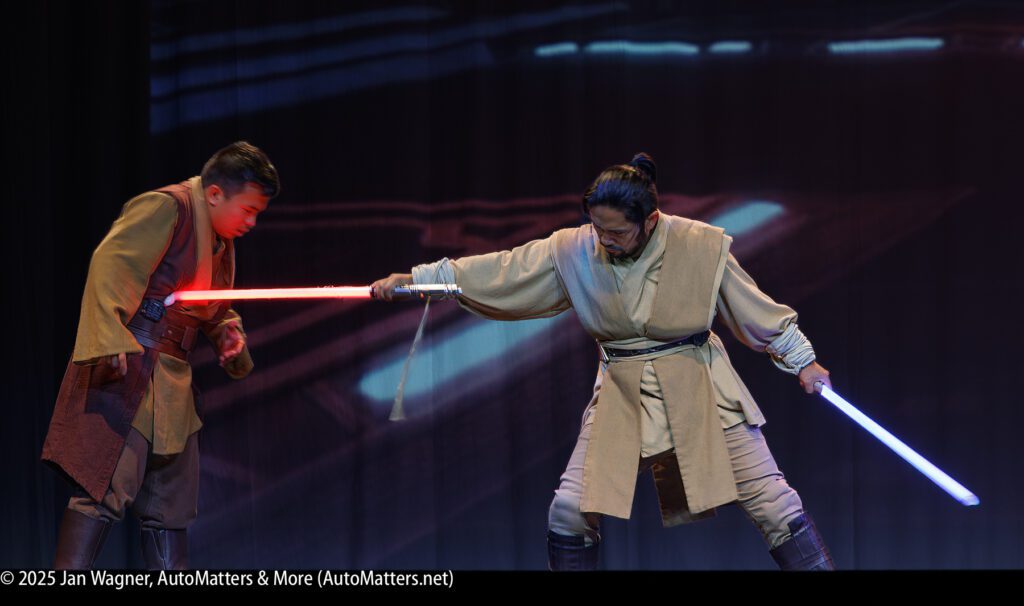 Two people in brown costumes perform a staged lightsaber duel with red and blue glowing swords on a dark stage.