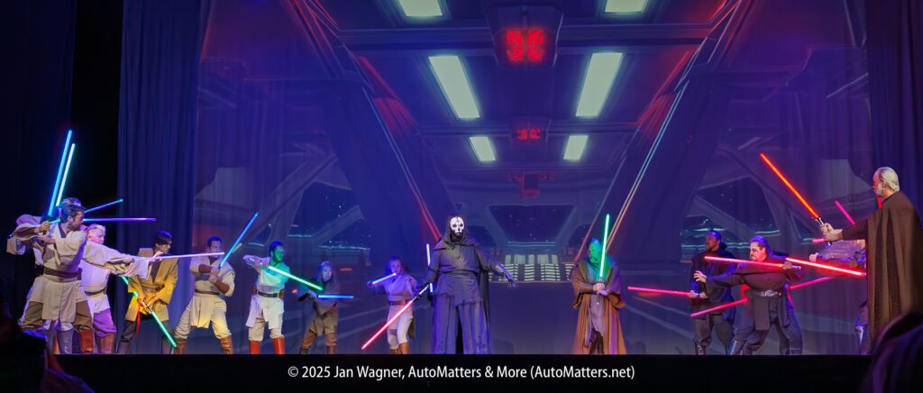 A group of people in Star Wars costumes, wielding lightsabers, confront a central masked figure on a stage with a sci-fi background.