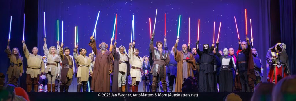 A large group of people in Star Wars costumes stand on stage holding lit lightsabers above their heads against a purple starry backdrop.