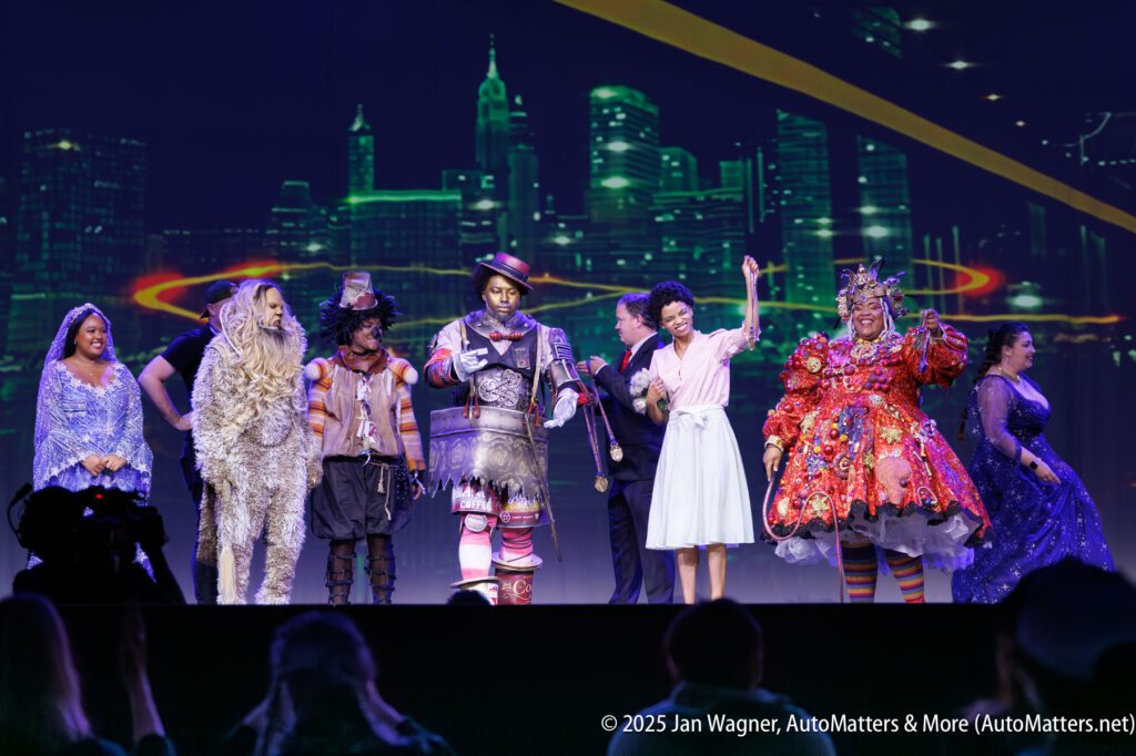A group of performers in colorful costumes stands on stage, posing for the audience, with a city skyline backdrop behind them.