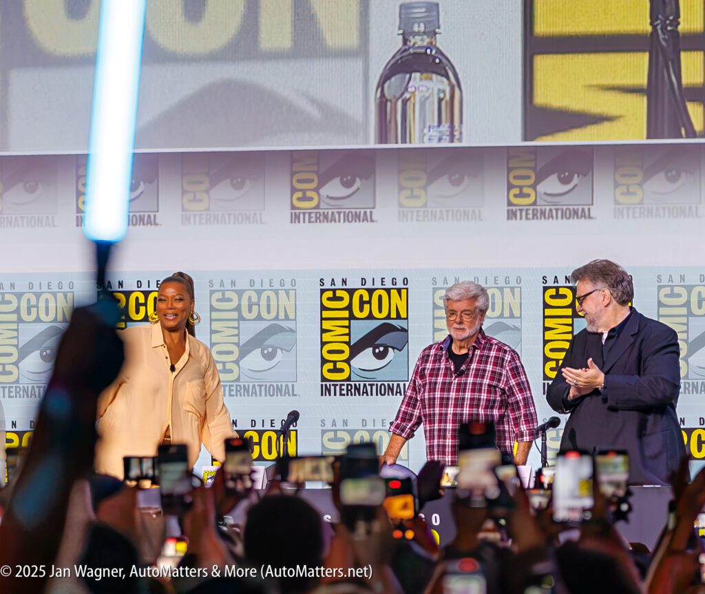 Three people stand on stage at San Diego Comic-Con International with a crowd taking photos and a lightsaber visible in the foreground. Comic-Con banners are displayed behind them.