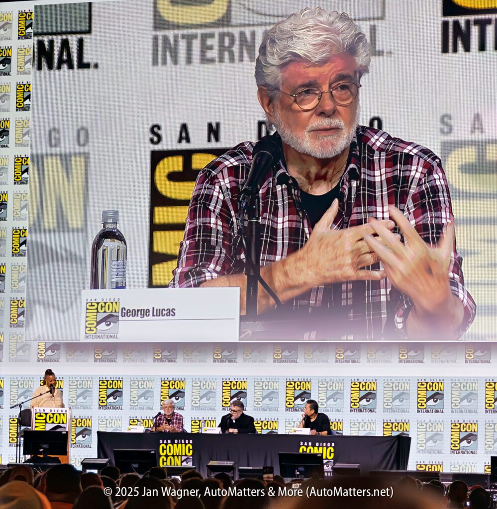 George Lucas speaks at a Comic-Con International panel, seated behind a nameplate, with other panelists and the event logo visible in the background.