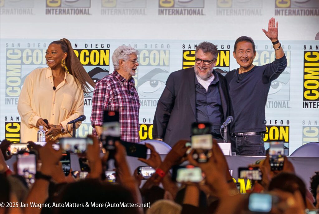 Four people stand on stage at San Diego Comic-Con International, with one person waving to the audience as attendees take photos.