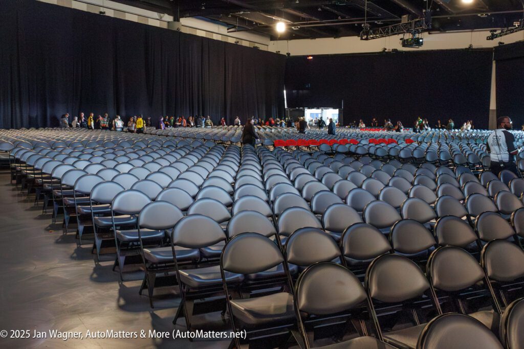 Rows of empty gray chairs fill a large indoor venue, with people entering and gathering near the far entrance at the back of the room.