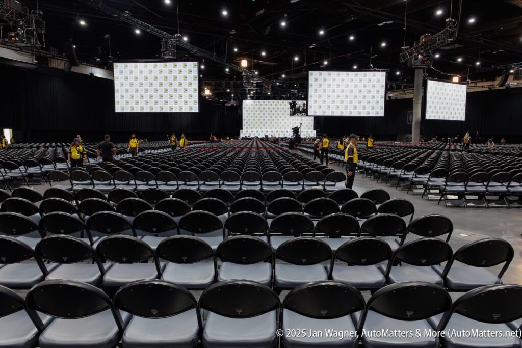 Rows of empty chairs face large screens and a stage in a convention hall, with staff in yellow vests standing throughout the space.