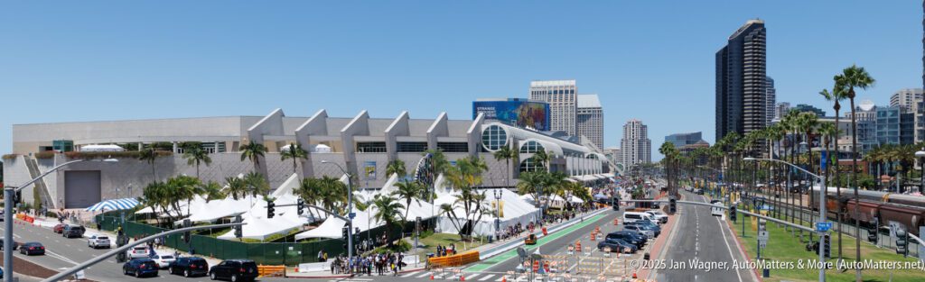 Wide view of a busy street lined with palm trees and tents beside a large modern building, with high-rise buildings in the background under a clear blue sky.