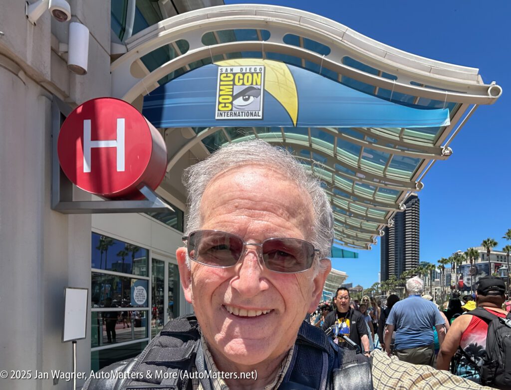 A man takes a selfie outside a building with a "Comic-Con International" banner; people are walking in the background under a clear blue sky.