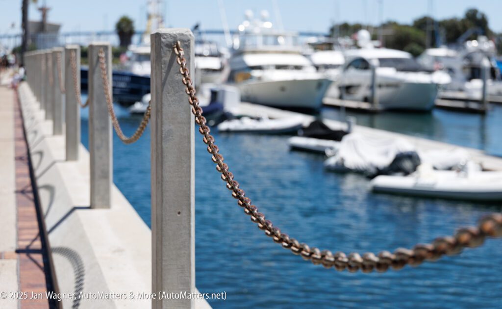 A metal chain runs between concrete posts along a marina walkway, with several boats docked on the water in the background.