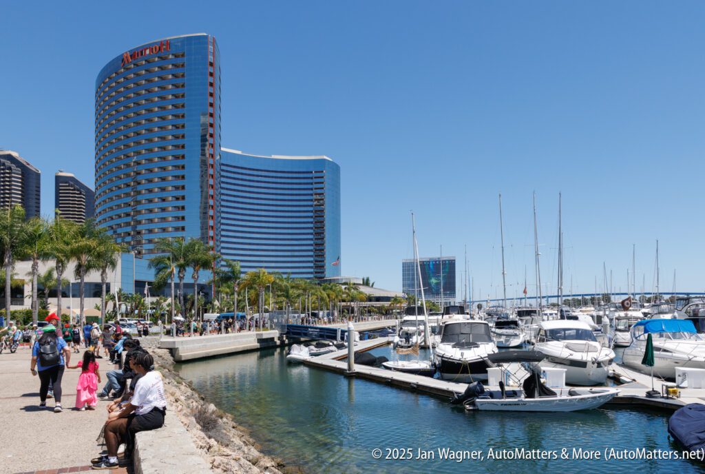People walk and sit along a waterfront marina with docked boats, palm trees, and a modern high-rise hotel under a clear blue sky.
