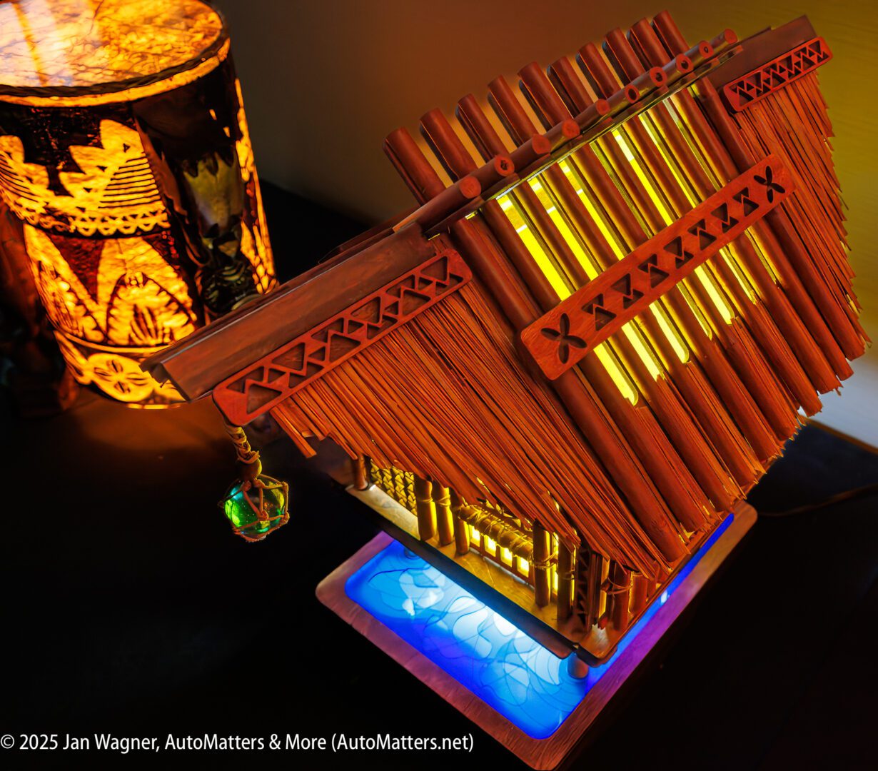 A model bamboo house with illuminated windows sits on a decorative base next to a lit, patterned lamp.