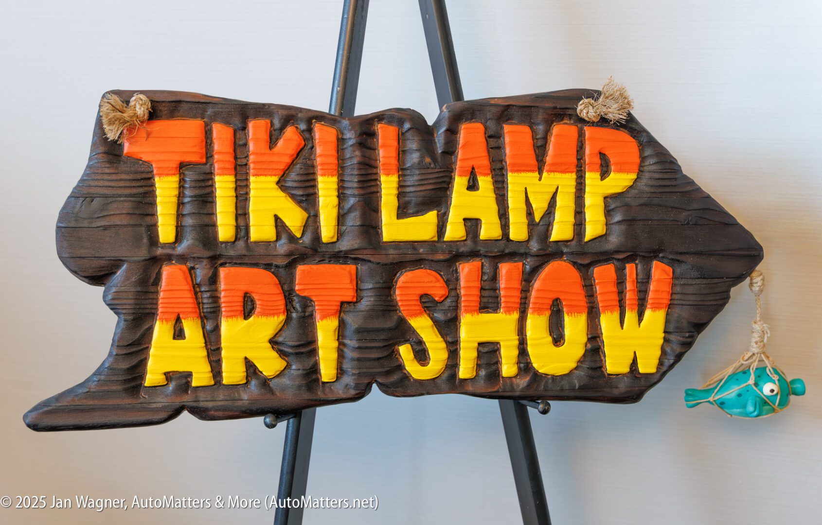 A wooden sign reads "Tiki Lamp Art Show" in bold yellow and orange letters, with a small blue fish decoration hanging on the right side.