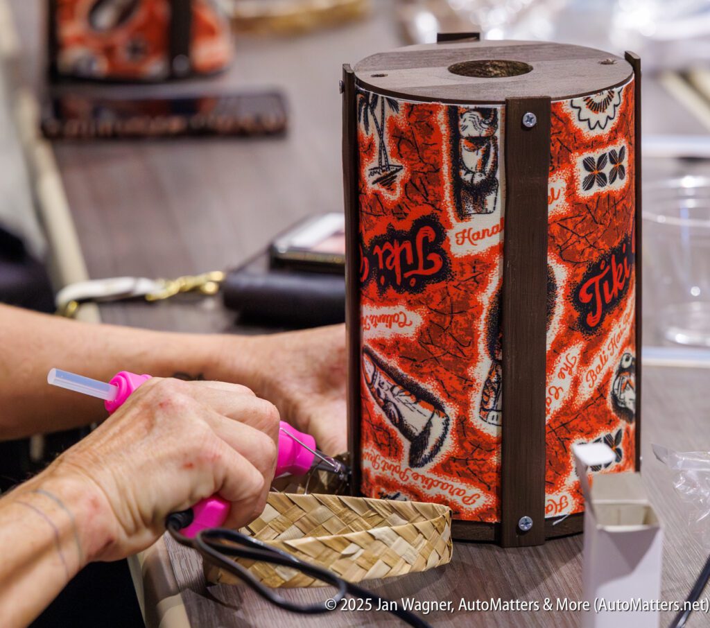 A person uses a pink hot glue gun to attach material to a small woven basket next to a Tiki-themed lamp on a table.