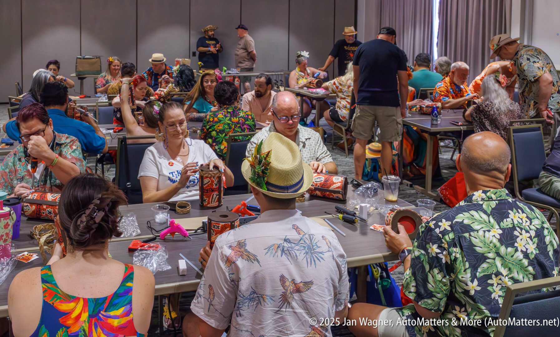 People sit at tables in a conference room, assembling items and wearing tropical or Hawaiian-themed clothing. Various craft materials are scattered on the tables.