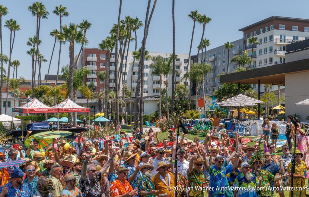 A large group of people in colorful Hawaiian shirts raises their arms at an outdoor event, with palm trees and apartment buildings in the background.