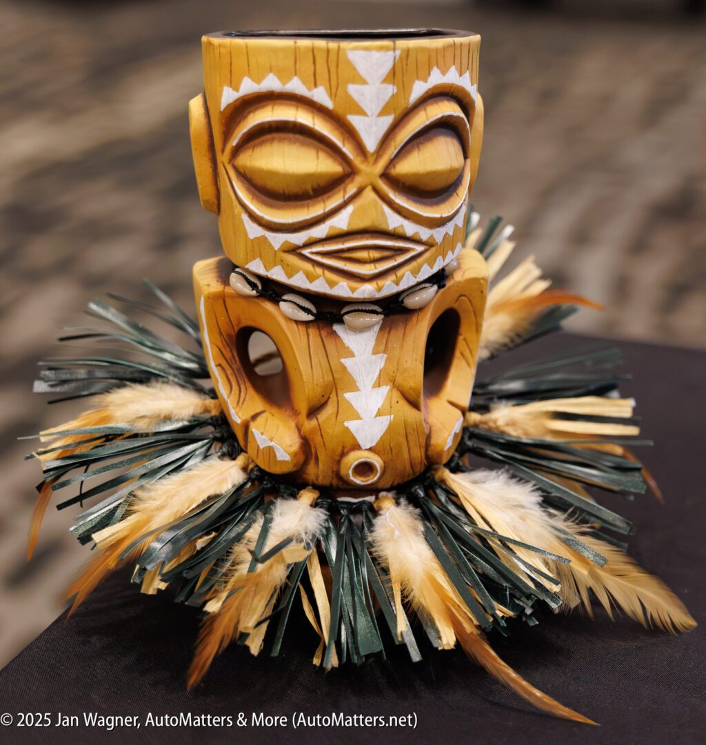 A carved wooden tiki mug with white tribal designs sits atop a circular base adorned with feathers and green leaves.