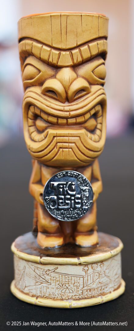 A carved tiki mug with a wide grin holds a metallic emblem reading "Tiki Oasis," standing on a round decorative base.