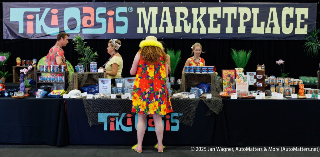 A woman in a yellow hat stands at a Tiki Oasis Marketplace booth, staffed by three people and displaying various products under a large sign.