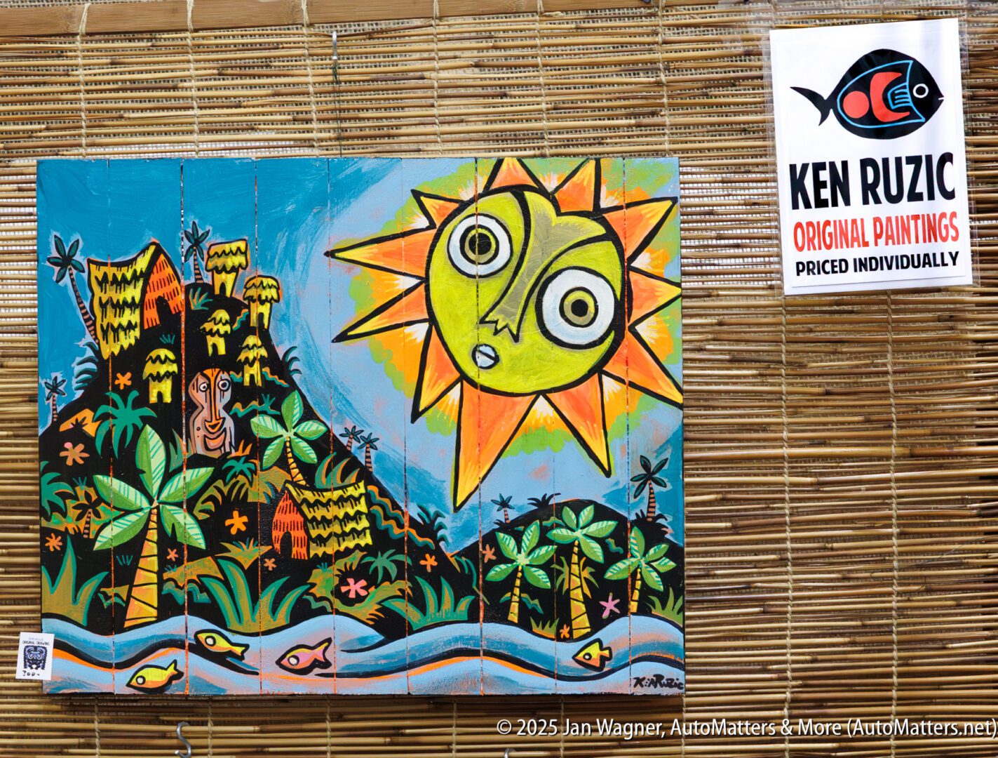 A colorful painting of a stylized sun, tropical huts, and fish is displayed on a bamboo mat, with a sign for Ken Ruzic original paintings hanging nearby.