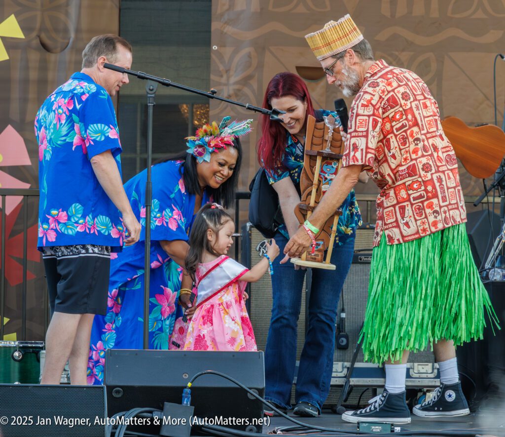 A young girl receives a trophy from a man in a grass skirt on stage, surrounded by three adults in colorful Hawaiian attire.