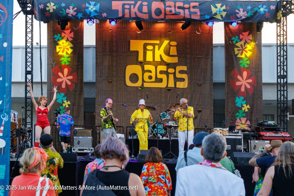 A band dressed in colorful outfits performs on an outdoor stage with a "Tiki Oasis" sign while an audience watches.