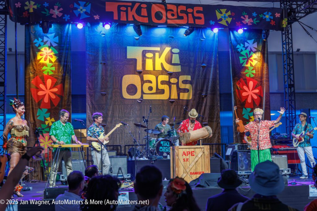 A band performs on stage at the Tiki Oasis event, with colorful decorations, instruments, and an audience watching.
