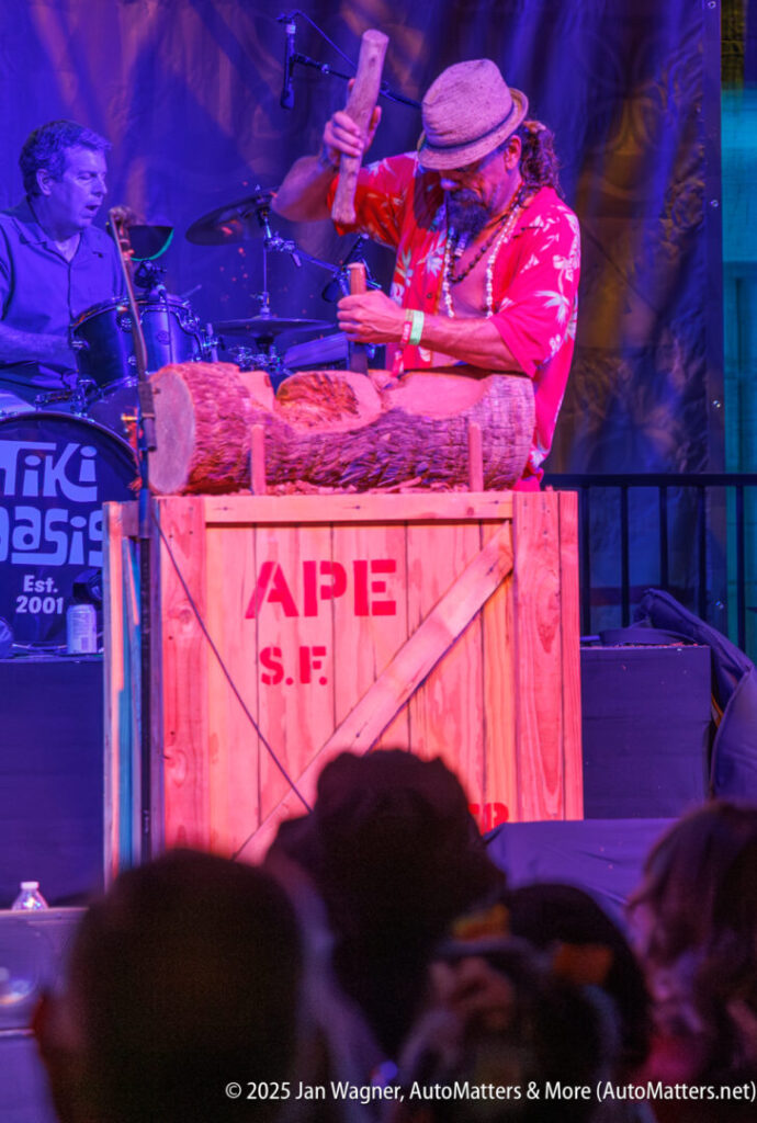 A musician in a red shirt and hat plays wooden log drums on a crate labeled "APE S.F." on a colorful stage, with a drummer visible in the background.