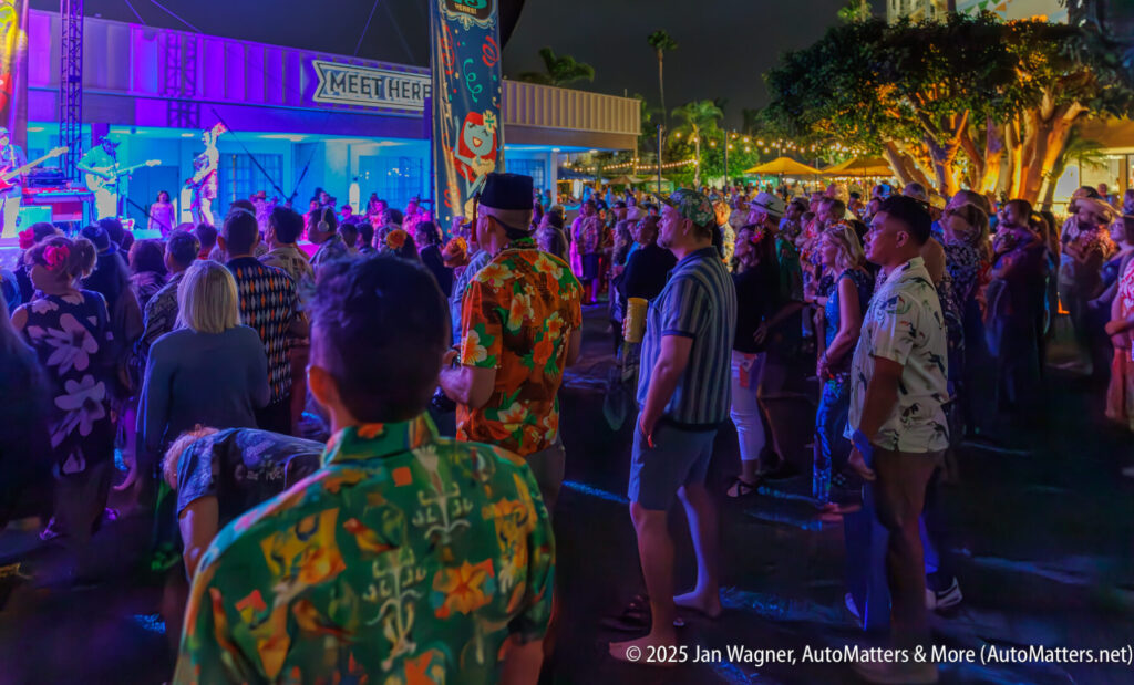 A crowd of people in Hawaiian shirts watch a nighttime outdoor event with colorful stage lighting.