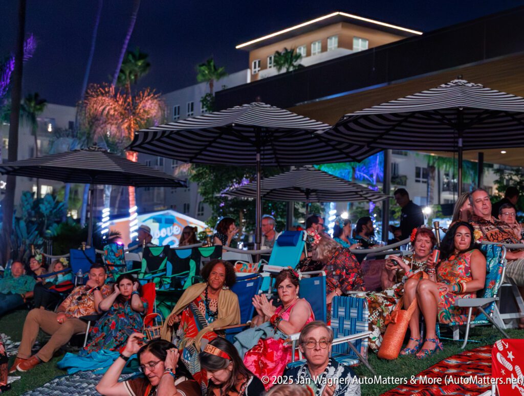 A group of people sits and reclines on lawn chairs and blankets under striped umbrellas at an outdoor nighttime event, with buildings and palm trees in the background.