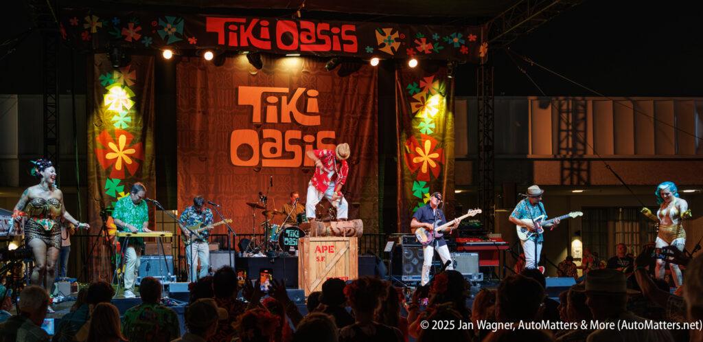 A band performs on stage at Tiki Oasis, with colorful tropical decorations and an audience watching at night.