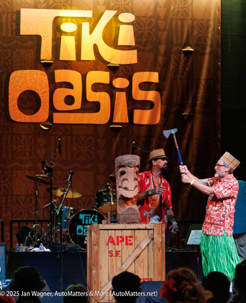 Two men stand on stage at a Tiki Oasis event; one man uses a mallet on a tiki statue atop a wooden crate labeled "APE S.F." Drums and a large Tiki Oasis sign are in the background.