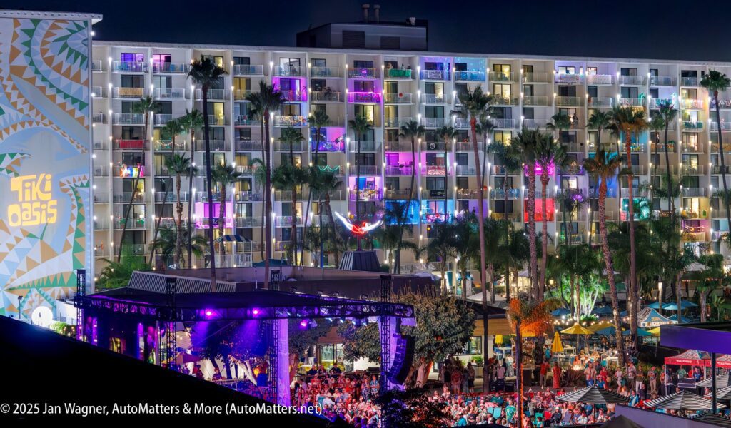 A large hotel building lit with colorful lights at night, palm trees in front, and a crowd gathered at an outdoor event with a stage.