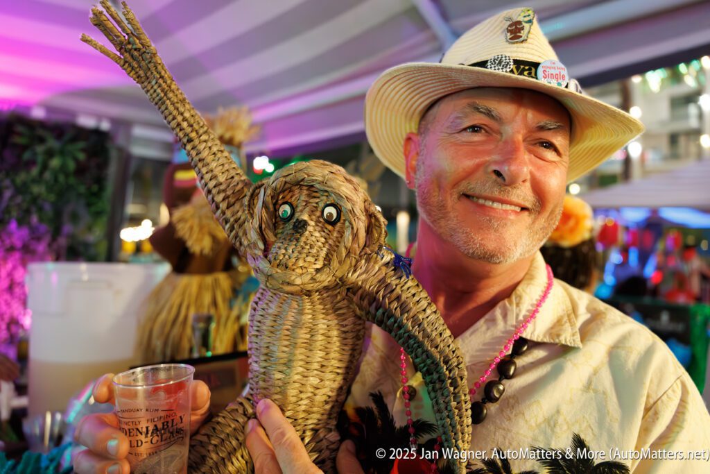 A man in a hat smiles while holding a woven straw monkey and a drink at a colorful event or festival.