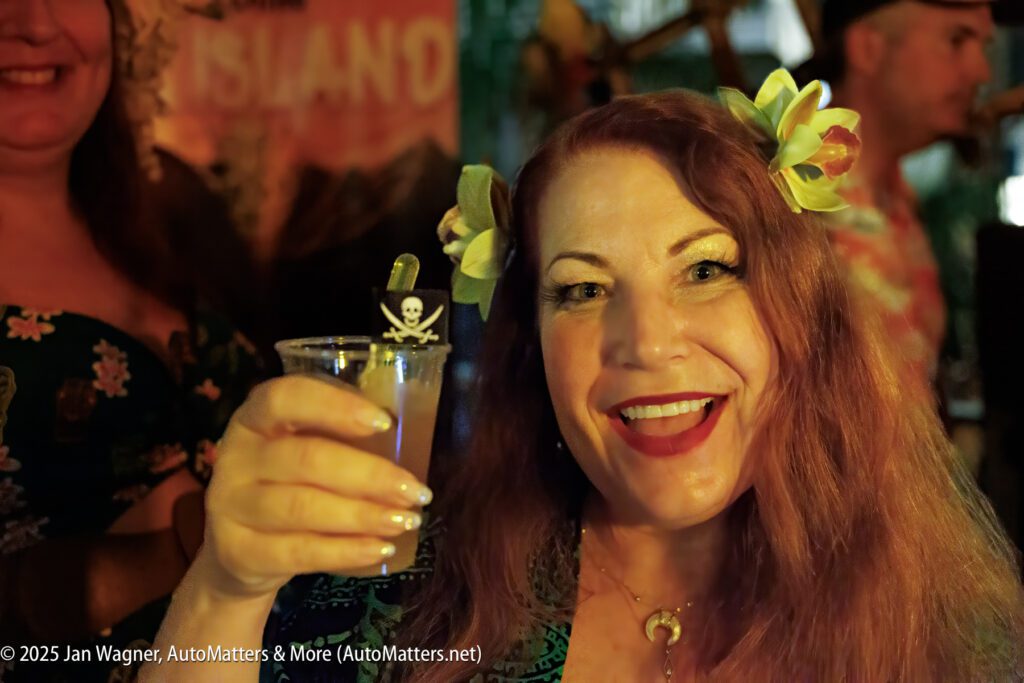 A smiling woman wearing flower hair clips holds a drink with a skull-and-crossbones stir stick at a festive indoor event.