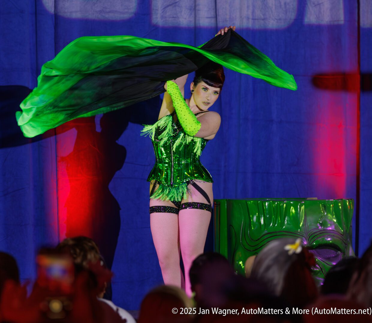 A performer in a green corset and gloves poses on stage with a large green fabric, in front of an audience and a blue backdrop.
