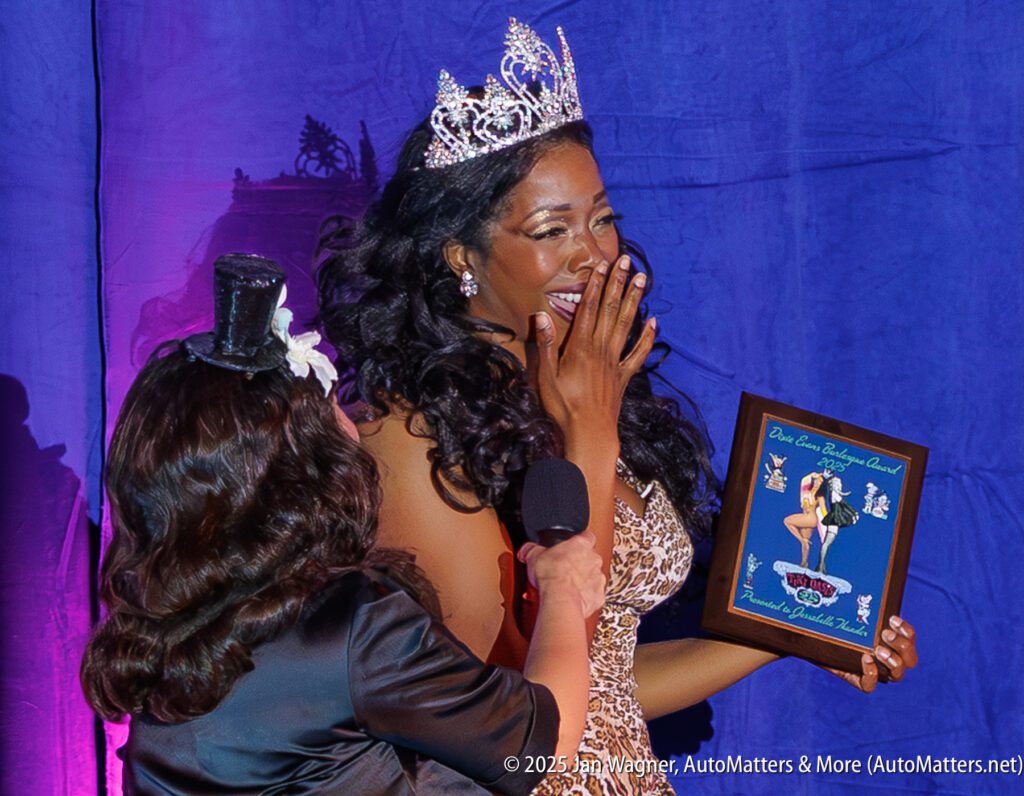 A woman in a crown and sequined dress holds a plaque and covers her mouth while another person with a microphone stands next to her against a purple backdrop.