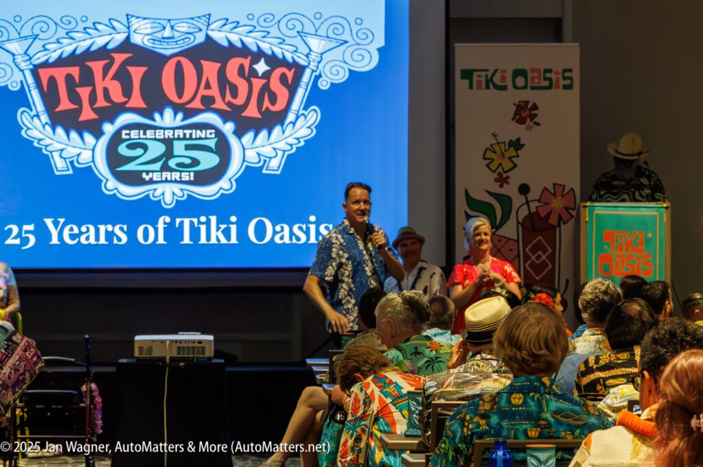A man speaks on stage at the Tiki Oasis 25th anniversary event, with an audience in Hawaiian shirts and event-themed decorations visible.