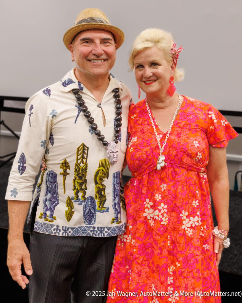 A man in a patterned shirt and hat stands next to a woman in a bright floral dress and shell necklace, both smiling at the camera.