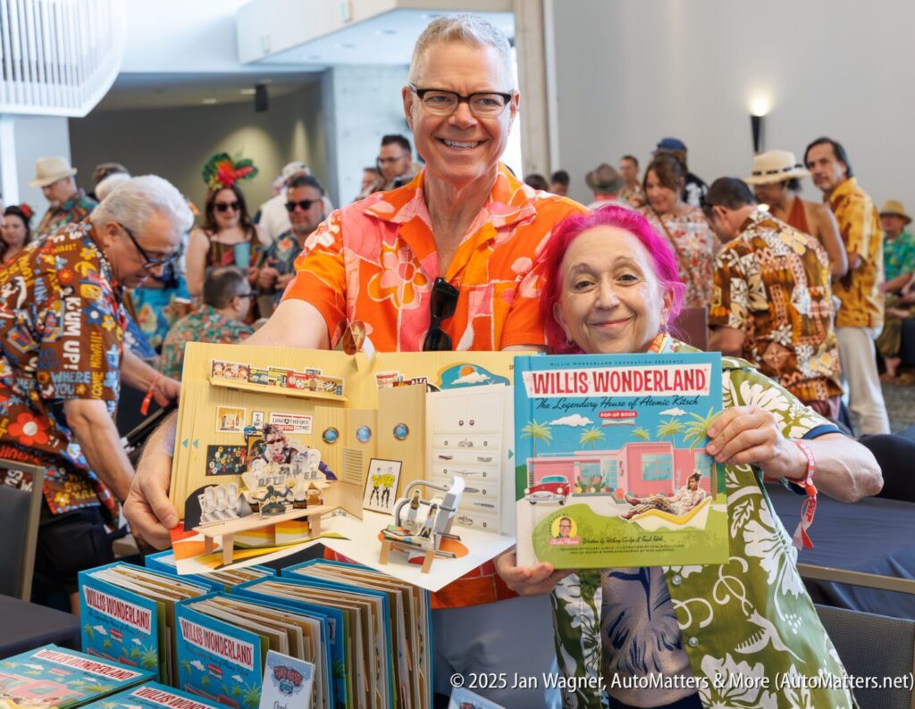 Two people in colorful shirts hold up copies of the book "Willis Wonderland" at a signing event. Other attendees are visible in the background.