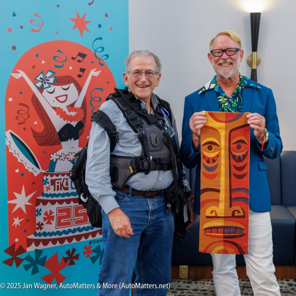 Two smiling men pose indoors; one holds a Tiki-themed artwork, and the other wears a utility vest. A colorful celebratory poster is visible in the background.