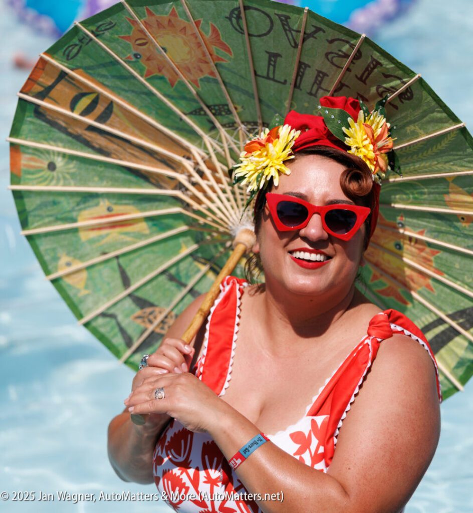 A woman in a red and white swimsuit and flower headband smiles while holding a decorative umbrella by a pool.