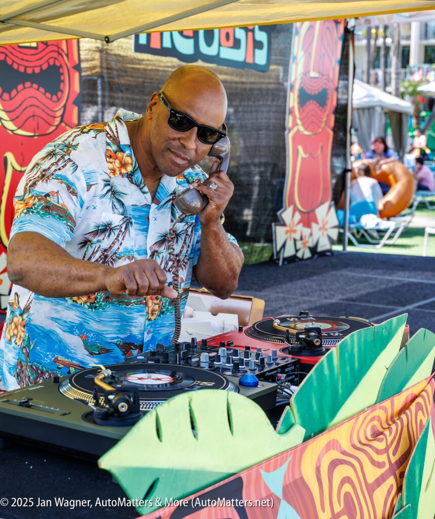 A man wearing sunglasses and a Hawaiian shirt operates DJ equipment at an outdoor event under a tent with colorful decorations.