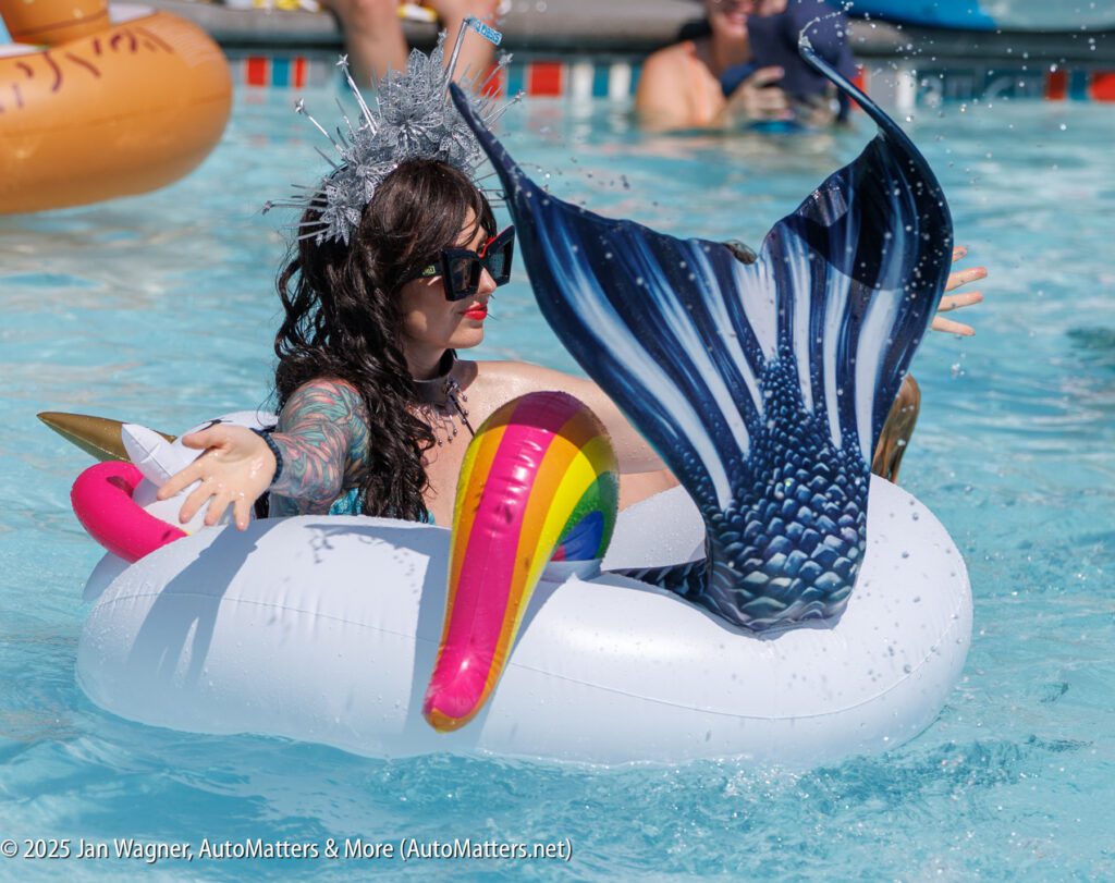 A woman wearing a crown and sunglasses floats in a pool on an inflatable unicorn, with a person in a mermaid tail in front of her.