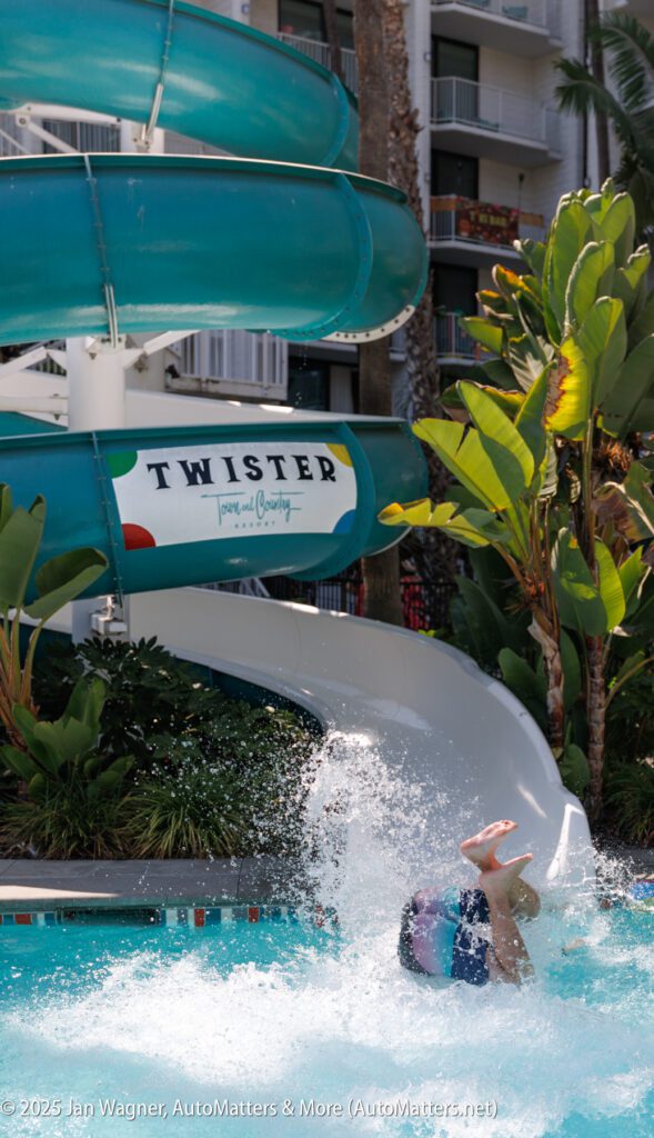 A person splashes headfirst into a pool at the bottom of a green water slide labeled "Twister" surrounded by tropical plants.