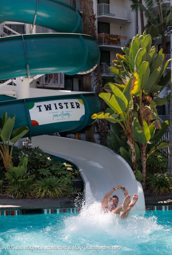 A person exits a green spiral water slide labeled "Twister" and splashes into a pool surrounded by tropical plants.