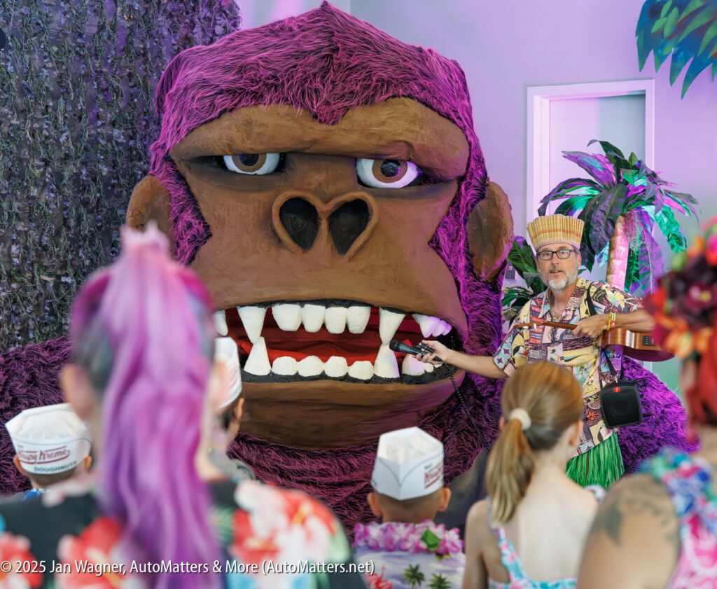 A man in a colorful outfit gestures toward a large purple gorilla statue while several people in paper hats watch in a brightly decorated room.