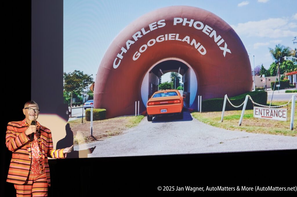 A person in a striped suit speaks on stage next to a projected image of a large donut-shaped structure labeled “Charles Phoenix GoogieLand” with a car driving through it.