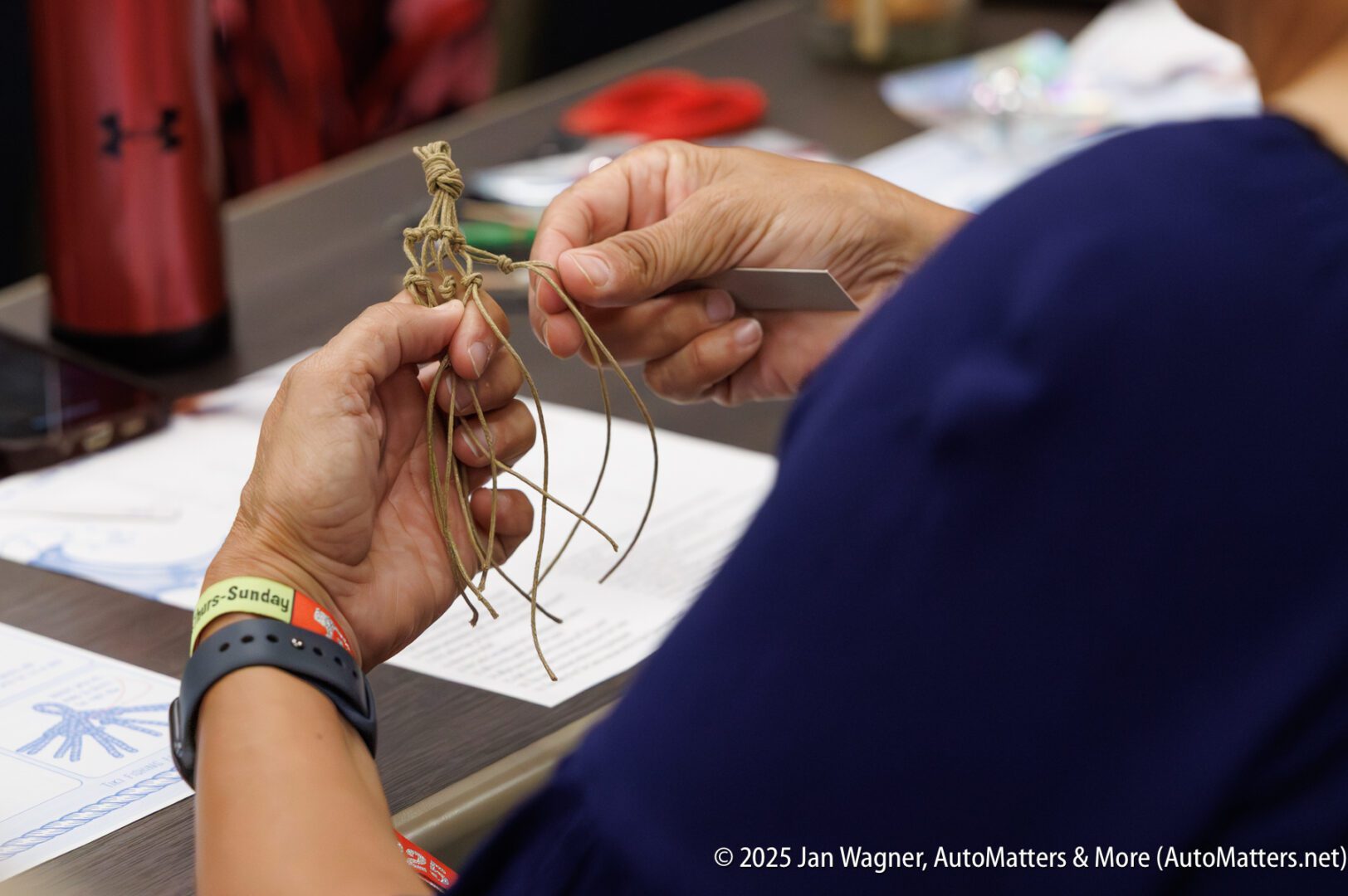 A person weaves a small figure out of twine at a table, holding several strands and using a green tool. Instructions and a red water bottle are visible nearby.