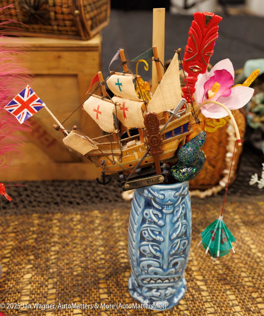 A decorative model sailing ship with small flags and flowers is arranged in a blue patterned vase on a wicker surface.