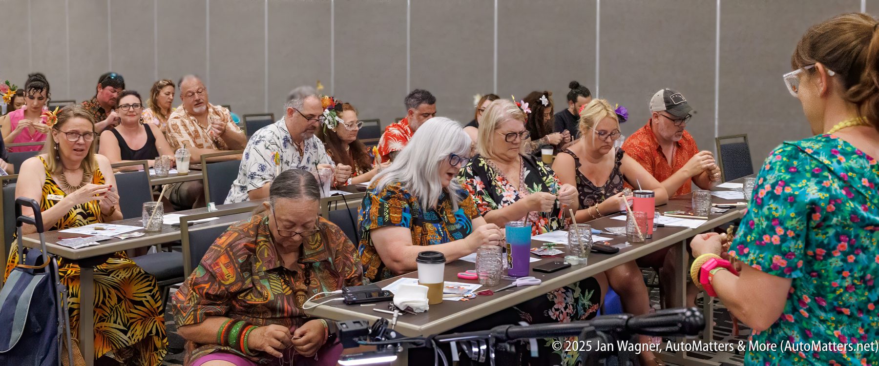 A group of adults, many wearing colorful floral clothing, participate in a hands-on activity at tables in a classroom setting.